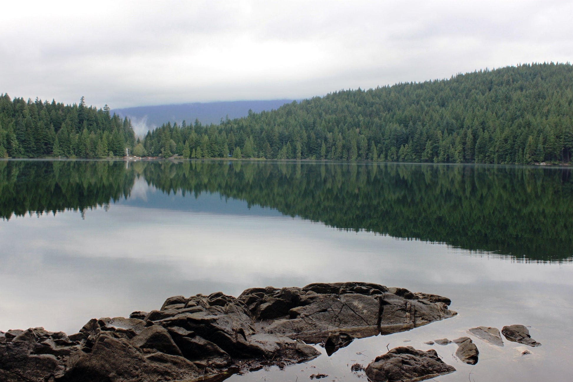 a rock in a lake surrounded by pine trees, overcast day, mist rising from a break in the hills across the lake.