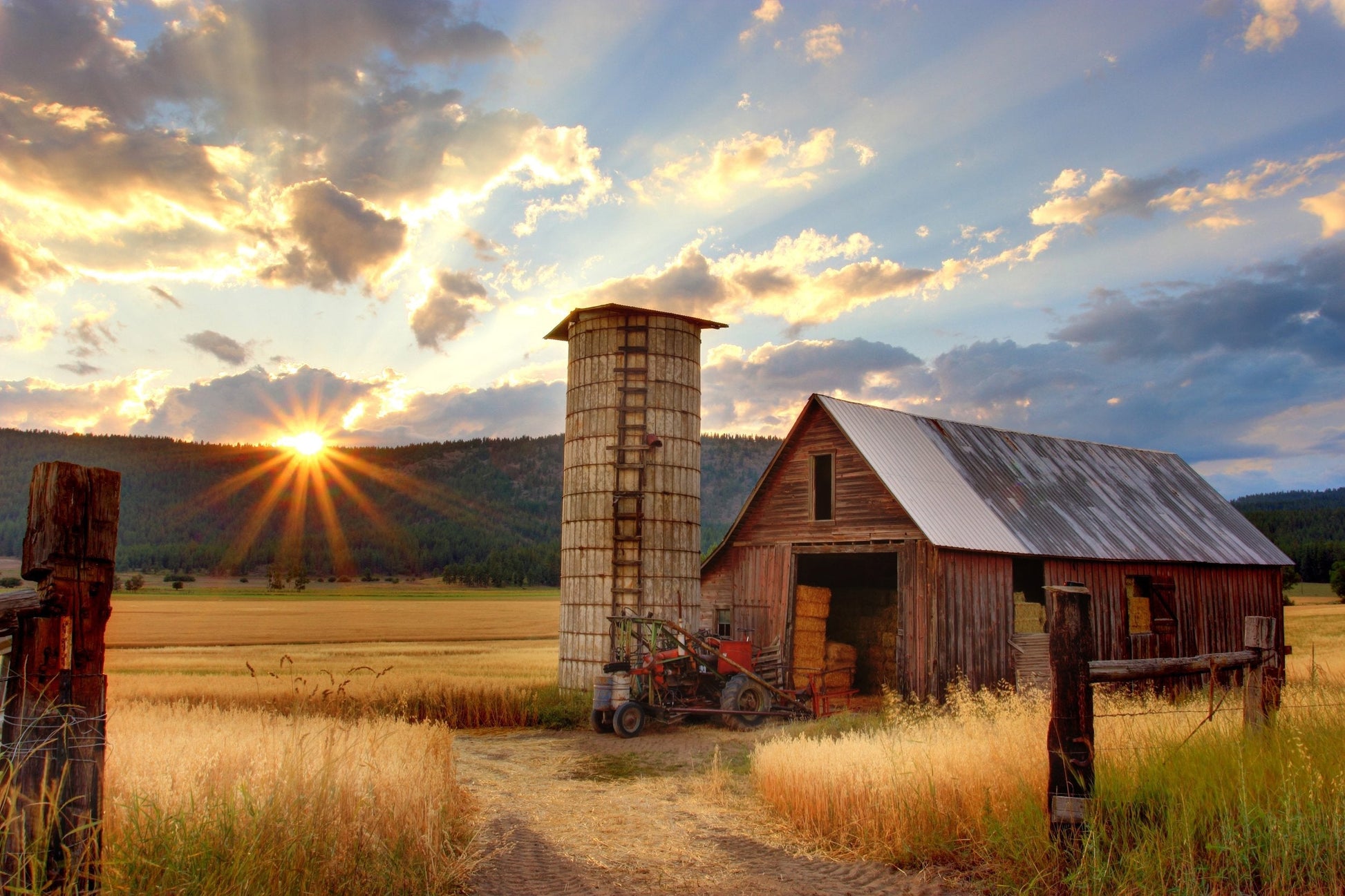 A hay barn in a field with a tractor in front of it. The sun sets over the hills in the backgound.