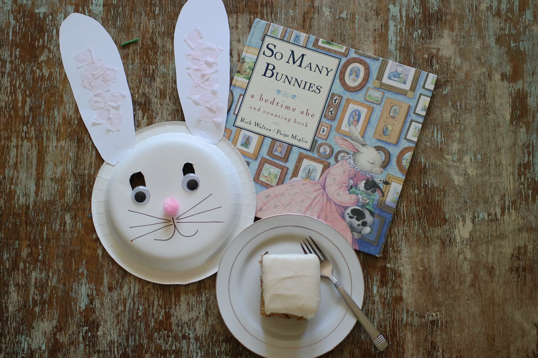 A homeschool project bunny mask, next too a piece of carrot cake and the book "So Many Bunnies" by Rick Walton and Paige Miglio.