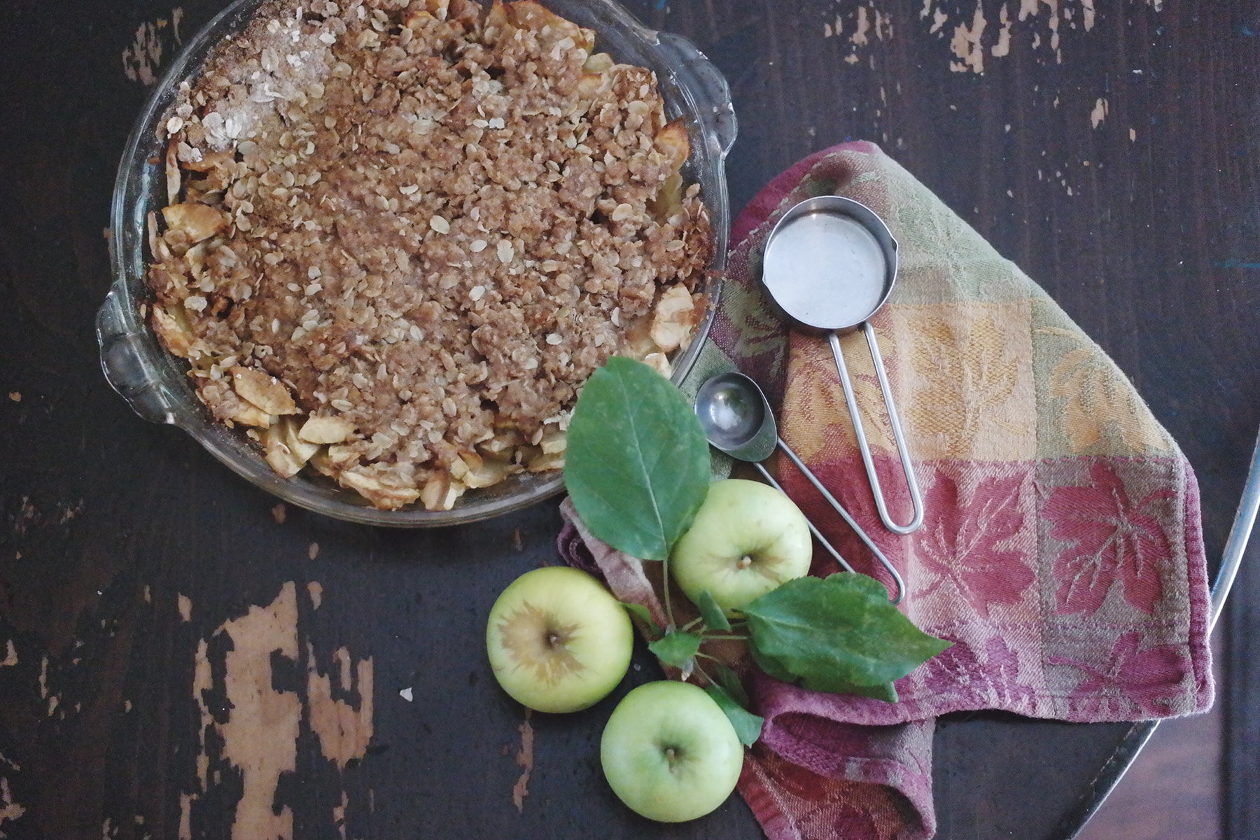 A homemade apple crumble next to some fresh apples, measuring utensils and a towel on a wooden table.
