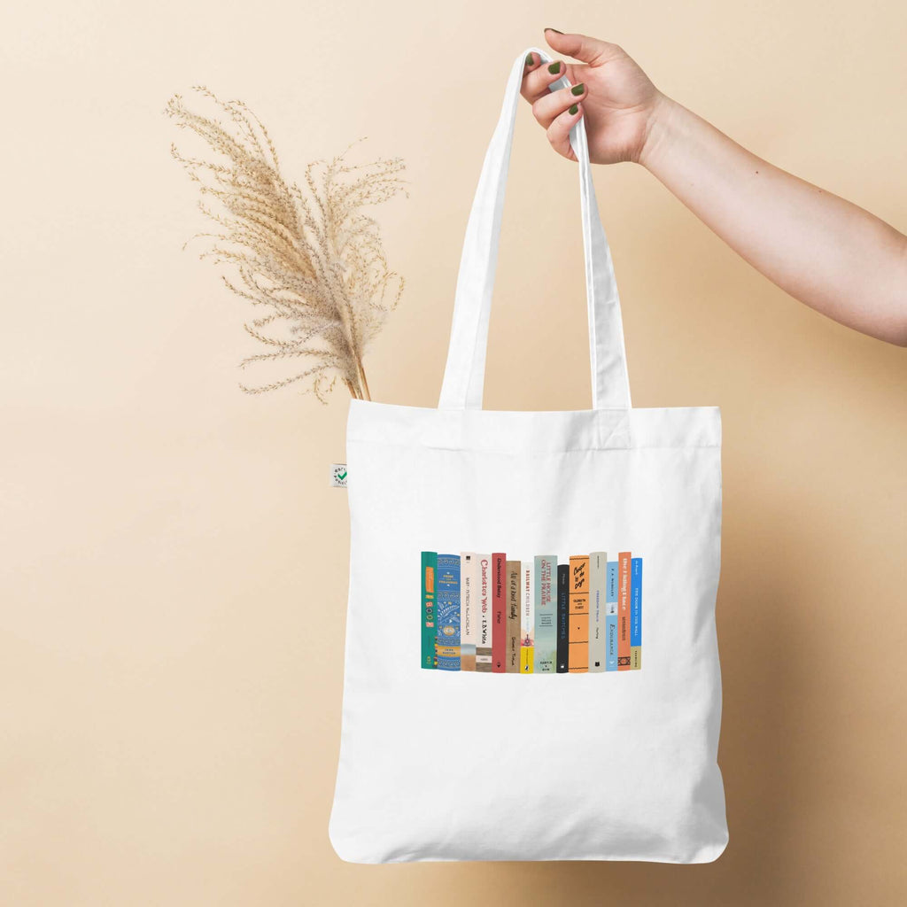 An image of a canvas tote bag held by a hand coming in from out of frame. The tote has an image of a painted bookshelf with books on it.