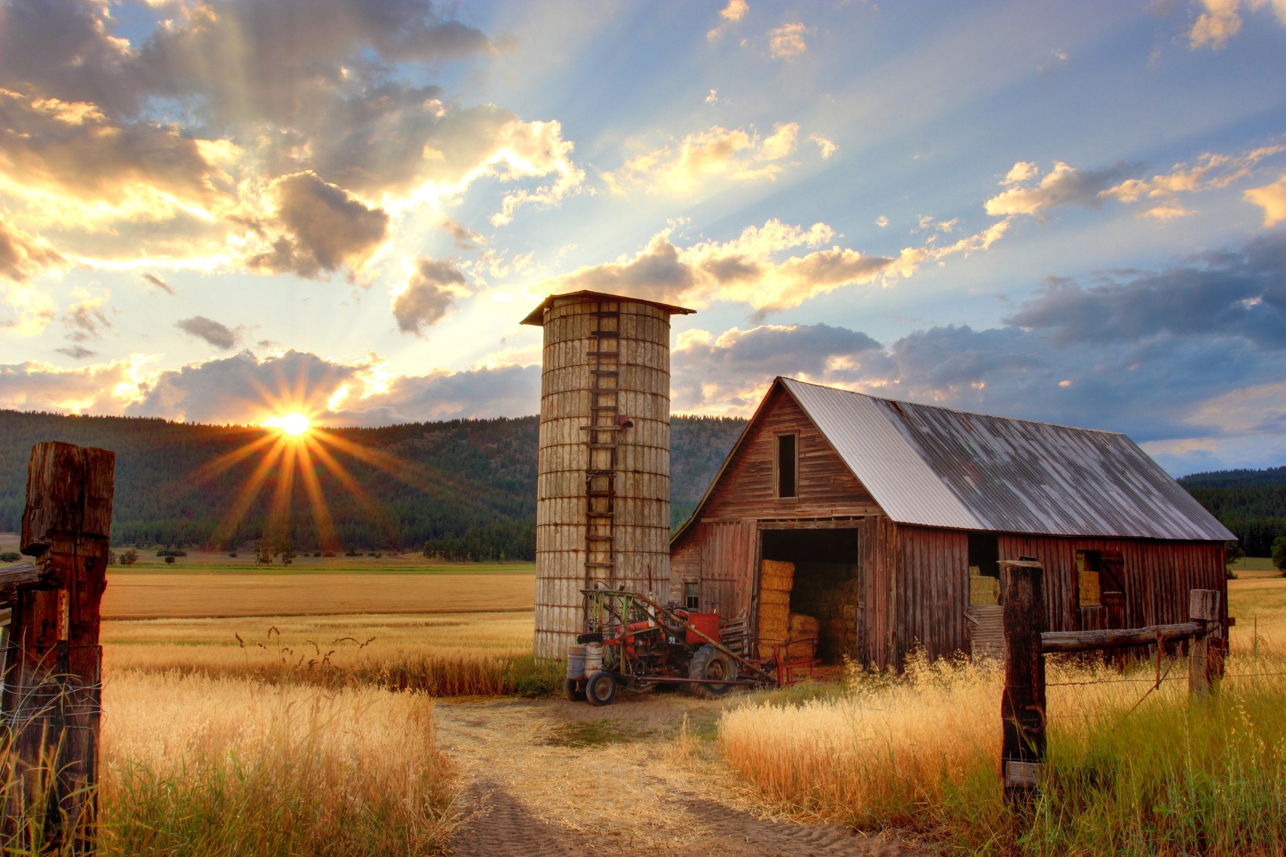 A hay barn in a field with a tractor in front of it. The sun sets over the hills in the backgound.